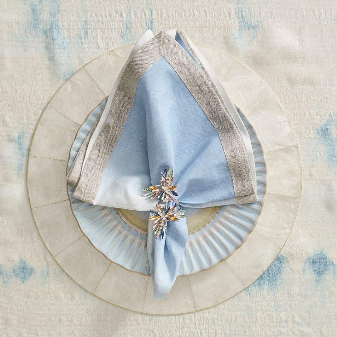 A pale blue linen napkin with a silver border, held by a jeweled floral napkin ring, rests on a scalloped light blue plate atop the Kim Seybert Capiz Shell Placemat Set 4 Natural, all arranged on a cream and blue tablecloth.