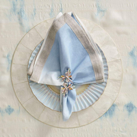 A pale blue linen napkin with a silver border, held by a jeweled floral napkin ring, rests on a scalloped light blue plate atop the Kim Seybert Capiz Shell Placemat Set 4 Natural, all arranged on a cream and blue tablecloth.