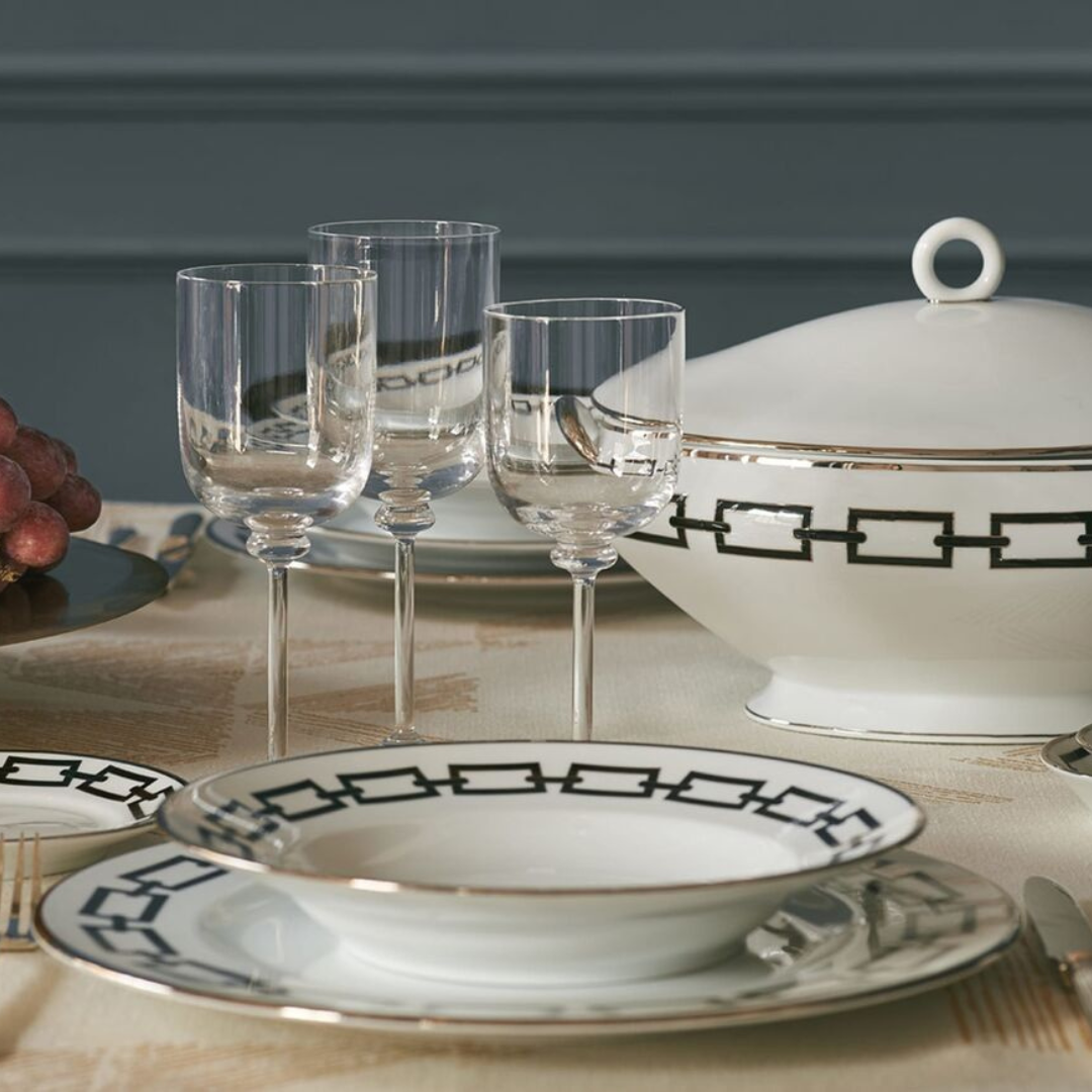 Elegant table setting with Ginori 1735 Catene Zaffiro dinnerware—designer porcelain plates with a black geometric border and gold trim. Includes three clear wine glasses, gold cutlery, and grapes on a beige tablecloth.