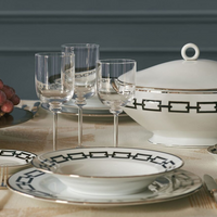 Elegant table setting with Ginori 1735 Catene Zaffiro dinnerware—designer porcelain plates with a black geometric border and gold trim. Includes three clear wine glasses, gold cutlery, and grapes on a beige tablecloth.