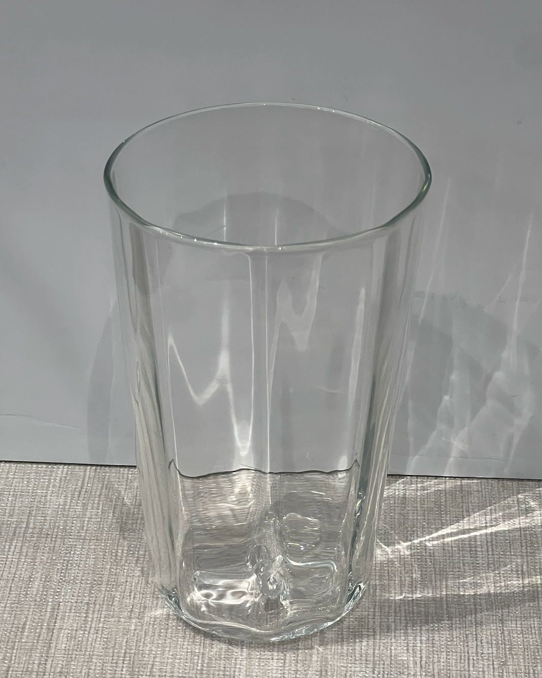 The Optic Everyday Cup Clear by Gary Bodker rests on a light, textured surface with a plain gray background. Light reflects off this handblown glass cup, casting subtle patterns and shadows.