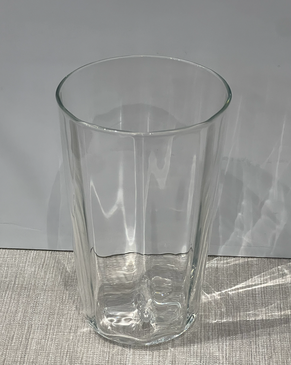 The Optic Everyday Cup Clear by Gary Bodker rests on a light, textured surface with a plain gray background. Light reflects off this handblown glass cup, casting subtle patterns and shadows.
