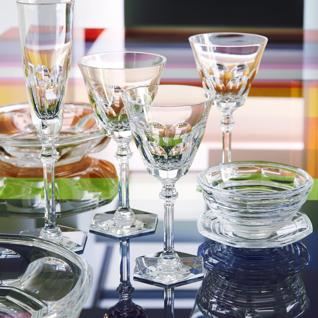 A set of elegant Baccarat Eve Wine Glasses with intricate designs and hexagonal bases is arranged on a reflective surface, accompanied by bowls and plates, against a colorful, blurred background.