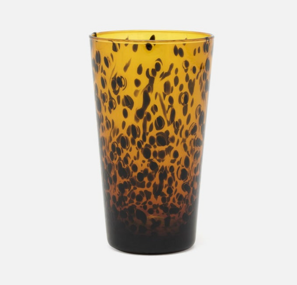 A tall highball glass from the Blue Pheasant Andrew Tortoise Drinkware set stands upright on a plain white background, its amber hand-blown glass adorned with a striking speckled pattern of dark brown spots reminiscent of classic tortoise shell. This elegant piece is part of a set of six highball glasses.