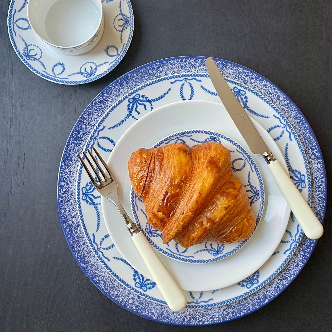 A golden croissant rests on a blue-patterned white plate beside Capdeco's Altea Flatware 5 Piece Setting with white handles, plus a matching teacup and saucer, all arranged elegantly on a dark table. The flatware is made in France.