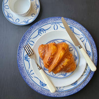A golden croissant rests on a blue-patterned white plate beside Capdeco's Altea Flatware 5 Piece Setting with white handles, plus a matching teacup and saucer, all arranged elegantly on a dark table. The flatware is made in France.