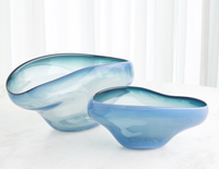 Two asymmetrical, translucent dark blue glass bowls of different sizes—part of the Harmony Bowl Dark Blue collection by Global Views / Surya—rest on a white surface with soft natural light filtering through a sheer white curtain behind them. Their elegant ombré design adds visual intrigue, making them a distinctive serving option for any contemporary table setting.