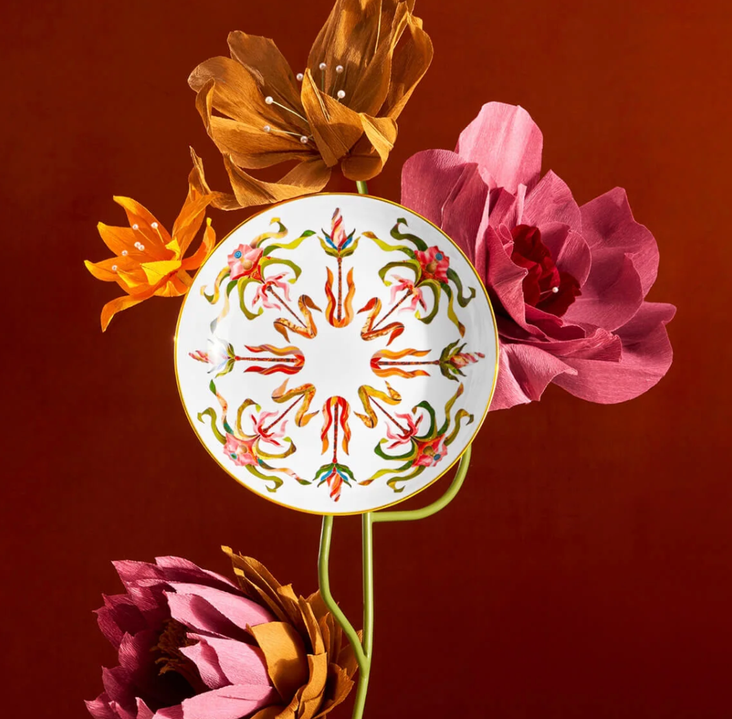 L’Objet Grand Tour Dinnerware round porcelain plate handcrafted in Portugal, featuring a symmetrical, colorful floral and leaf pattern with 24k gold accents, displayed among several large paper flowers on stems.