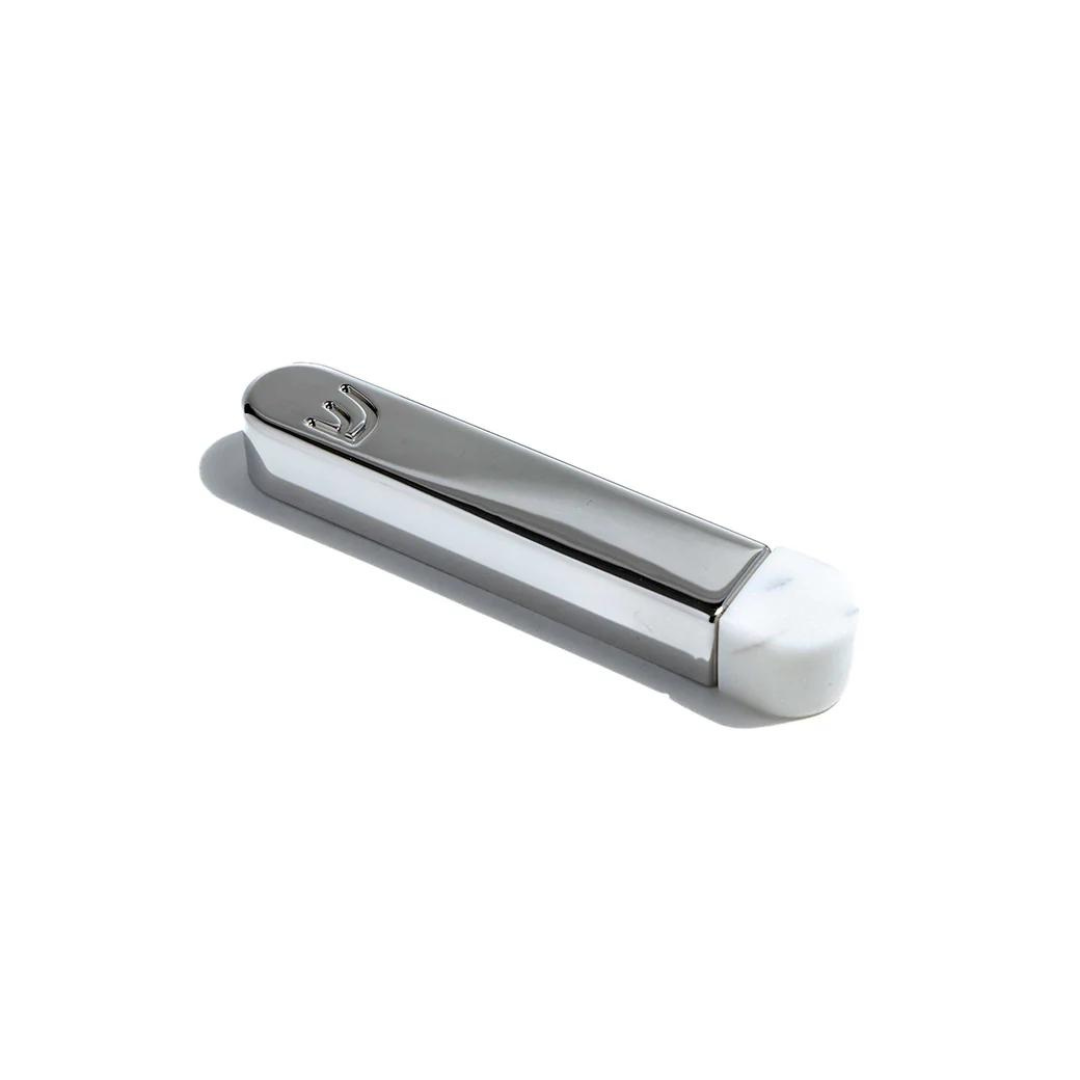 The Coluna Mezuzah Marble & Silver - Small by Anna New York features a silver case with a white marble base, engraved with a Hebrew letter symbolizing home protection. It rests on a white surface, casting a soft shadow.