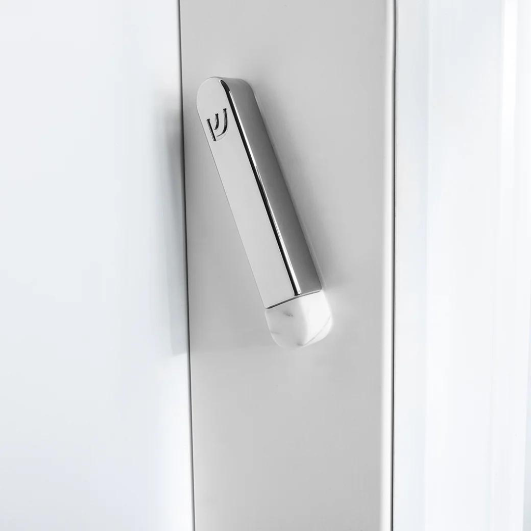 A sleek, metallic door handle with a small logo and a rounded, Carrara Marble-like white end is mounted vertically on a white door next to the Anna New York Coluna Mezuzah Marble & Silver - Small.