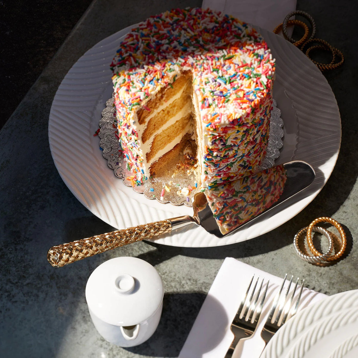 A tall, layered cake with colorful sprinkles sits on L'Objet's Corde White Dinnerware, next to a fine porcelain sugar bowl. A slice rests by a gold-handled server, with forks and napkins nearby.