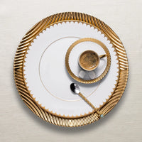 A small espresso cup from L'Objet's Corde Dinnerware Gold collection, with gold rim and matching saucer and large plate, sits alongside a gold-handled spoon on a textured off-white background.