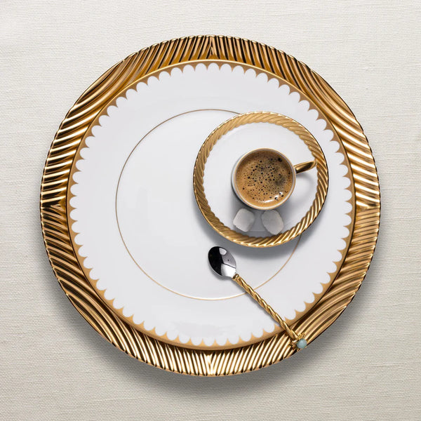 A small espresso cup from L'Objet's Corde Dinnerware Gold collection, with gold rim and matching saucer and large plate, sits alongside a gold-handled spoon on a textured off-white background.