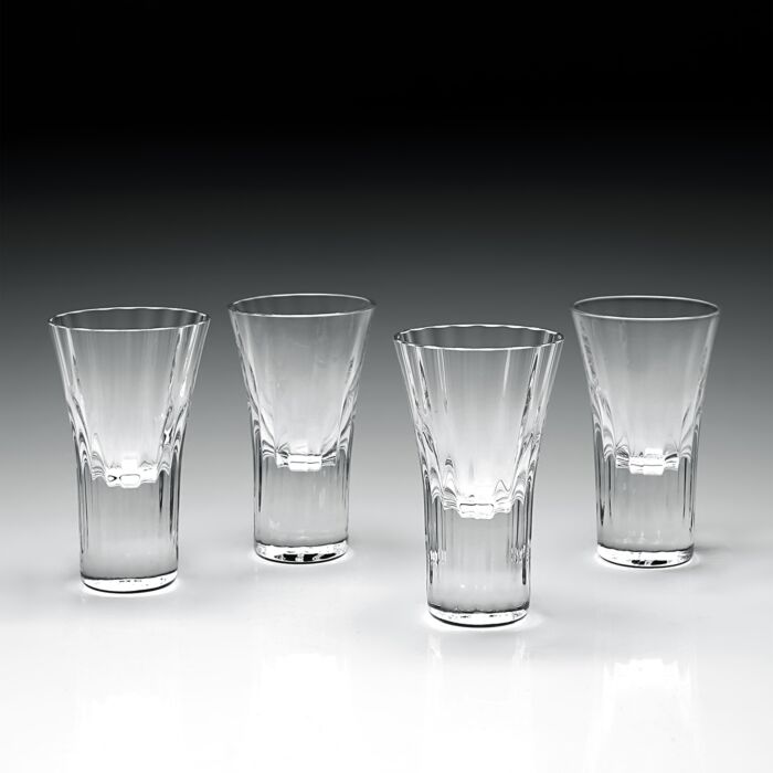 Four William Yeoward Corinne Shot Glasses—2 oz each, handmade crystal, set of 4—are lined up on a reflective surface with a gradient background transitioning from black above to light gray below.