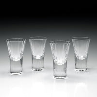 Four William Yeoward Corinne Shot Glasses—2 oz each, handmade crystal, set of 4—are lined up on a reflective surface with a gradient background transitioning from black above to light gray below.