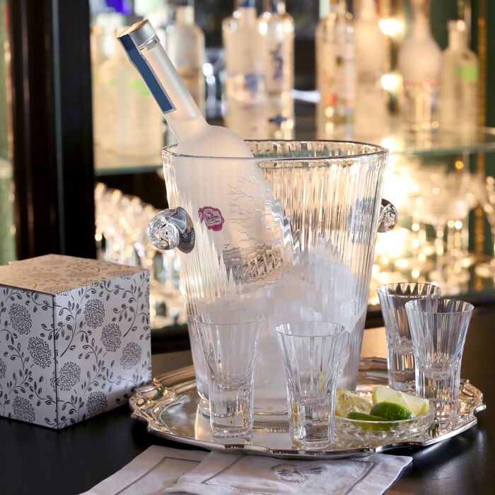 A bottle of vodka rests in a glass ice bucket on a silver tray with lime wedges, the William Yeoward Corinne Shot Glass Set of 4, and a decorative box displayed; glass bottles and barware blur in the background.