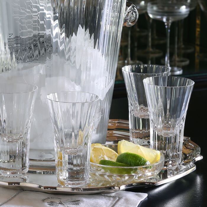 A silver tray holds four William Yeoward Corinne Shot Glasses (set of 4), each 2 oz handmade crystal glass, beside a glass ice bucket and a dish with lemon and lime wedges. Reflections of more glasses shimmer in the background.