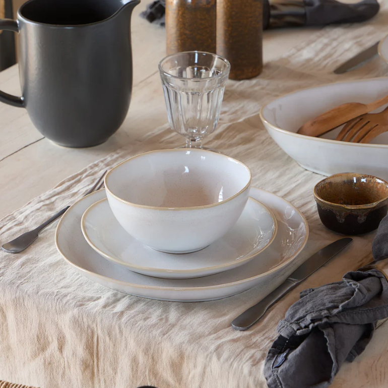 A neutral table setting features Casafina/Costa Nova Brisa Branca Dinnerware—a white stoneware bowl, two plates, silverware, a clear glass, black pitcher, and gray napkin on a beige tablecloth. Wooden serving utensils and more Brisa Branca pieces appear in the background.