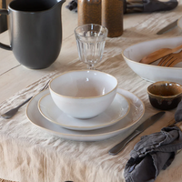 A neutral table setting features Casafina/Costa Nova Brisa Branca Dinnerware—a white stoneware bowl, two plates, silverware, a clear glass, black pitcher, and gray napkin on a beige tablecloth. Wooden serving utensils and more Brisa Branca pieces appear in the background.