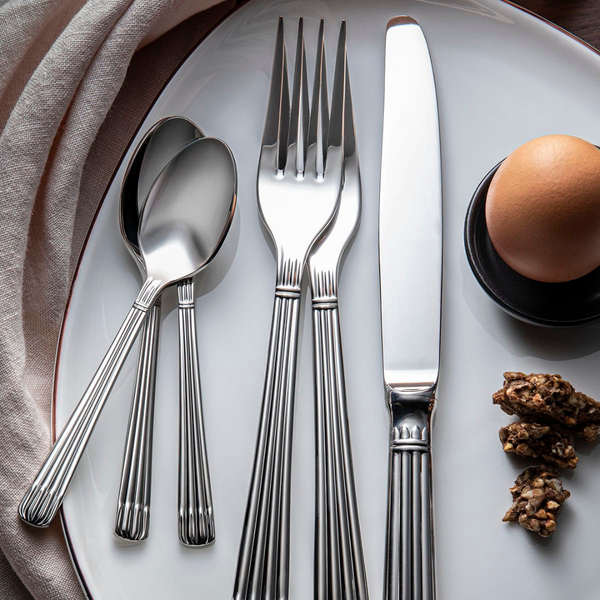 A white plate displays the Christofle Osiris Flatware 5 Piece Setting in stainless steel—two spoons, two forks, and a knife—alongside a brown egg in a holder, some granola, and a partially visible beige napkin.
