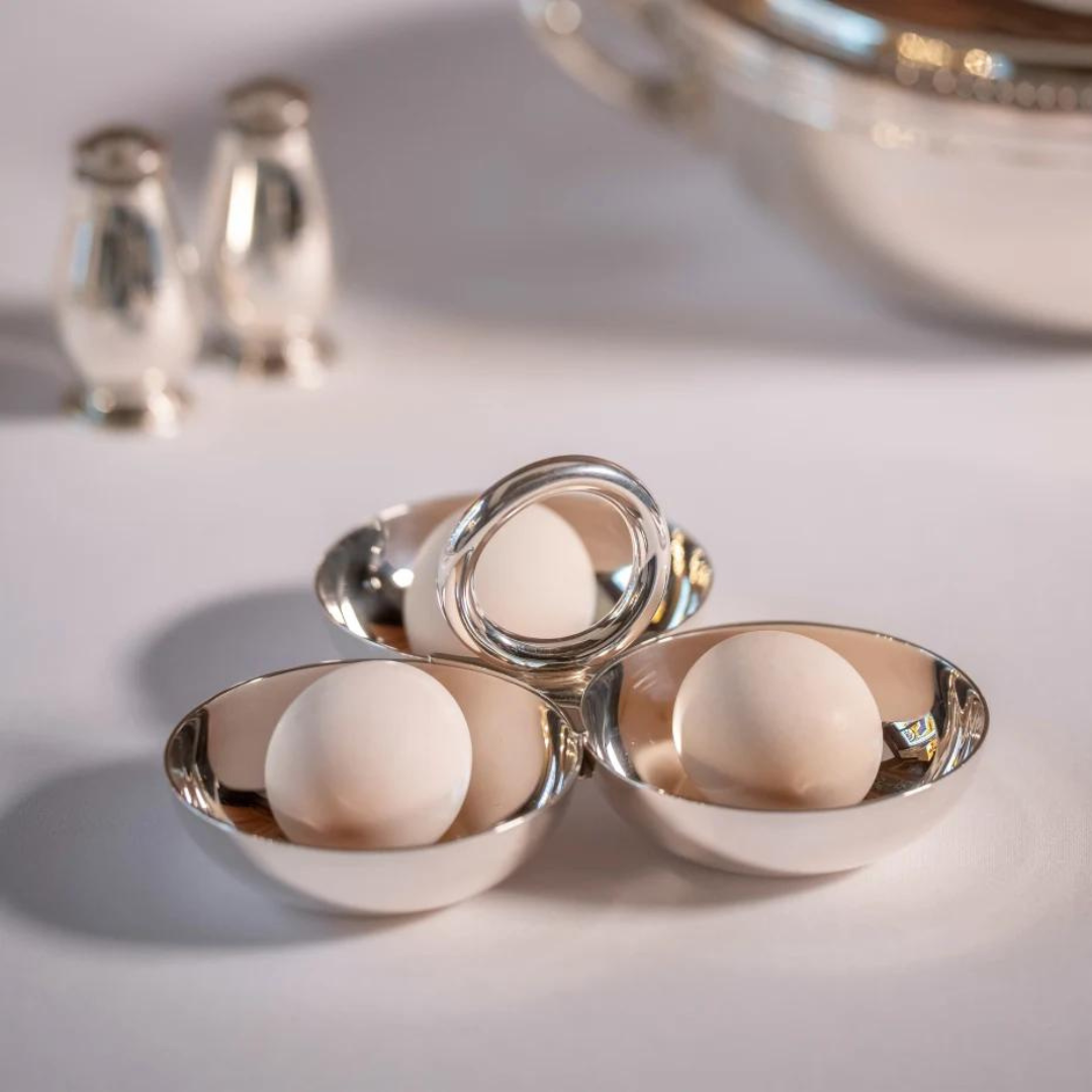 Three Christofle Vertigo Silver Plated 3 Part Servers, each with a white egg, are arranged in a triangle on a white surface. Blurred in the background are two silver shakers and a large serving dish from the Vertigo collection.