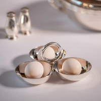 Three Christofle Vertigo Silver Plated 3 Part Servers, each with a white egg, are arranged in a triangle on a white surface. Blurred in the background are two silver shakers and a large serving dish from the Vertigo collection.