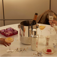 A festive celebration with cocktails, elegant glassware, an ashtray, and confetti features two hands holding drinks and the Christofle GRAPHIK Silver Plated Champagne Cooler as a stylish centerpiece.
