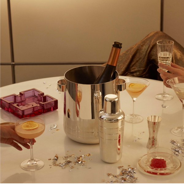 A festive celebration with cocktails, elegant glassware, an ashtray, and confetti features two hands holding drinks and the Christofle GRAPHIK Silver Plated Champagne Cooler as a stylish centerpiece.