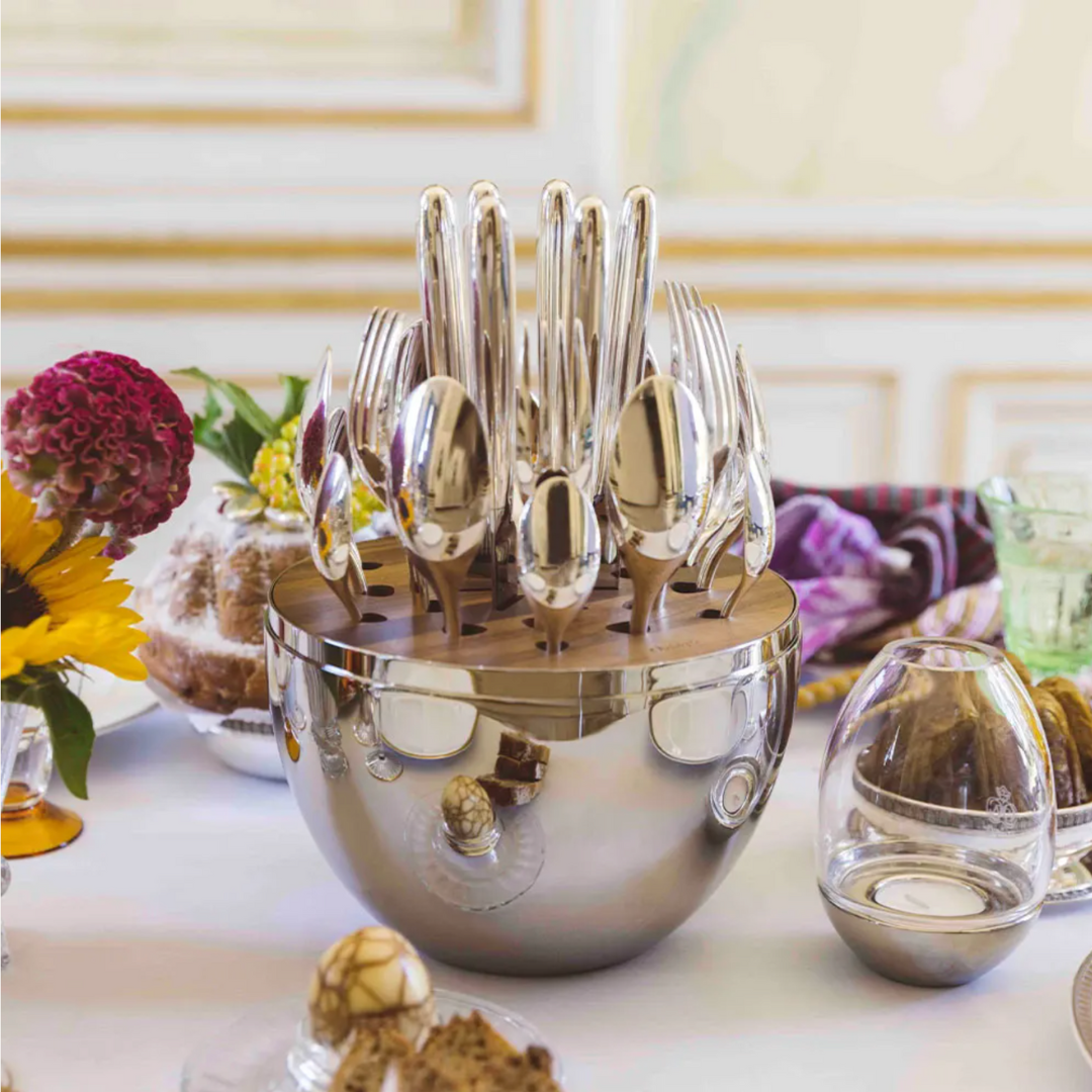 The Christofle Mood Flatware 24 Piece Set - Silver Plated is elegantly displayed in a polished silver egg on a white tablecloth, surrounded by flowers, a bundt cake, and a glass votive.