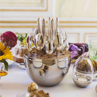 The Christofle Mood Flatware 24 Piece Set - Silver Plated is elegantly displayed in a polished silver egg on a white tablecloth, surrounded by flowers, a bundt cake, and a glass votive.