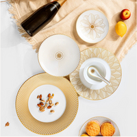 A flat lay of Christofle Malmaison Imperiale Porcelain Dinnerware Gold plates, a bowl with a spoon, cookies, nuts, lemon, apple, and a bottle arranged on beige fabric over a white surface.