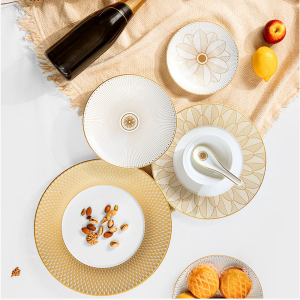 A flat lay of Christofle Malmaison Imperiale Porcelain Dinnerware Gold plates, a bowl with a spoon, cookies, nuts, lemon, apple, and a bottle arranged on beige fabric over a white surface.