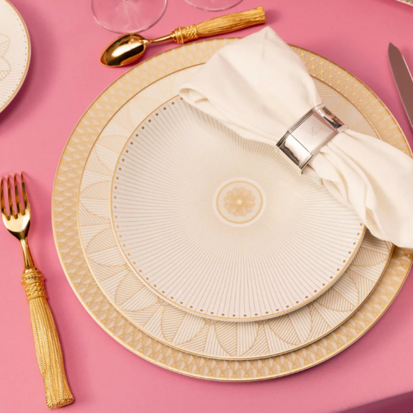 A formal table setting with a Christofle Malmaison Imperiale Porcelain Dinnerware Gold dinner plate, a folded white napkin in a silver ring, and gold utensils on a pink tablecloth.