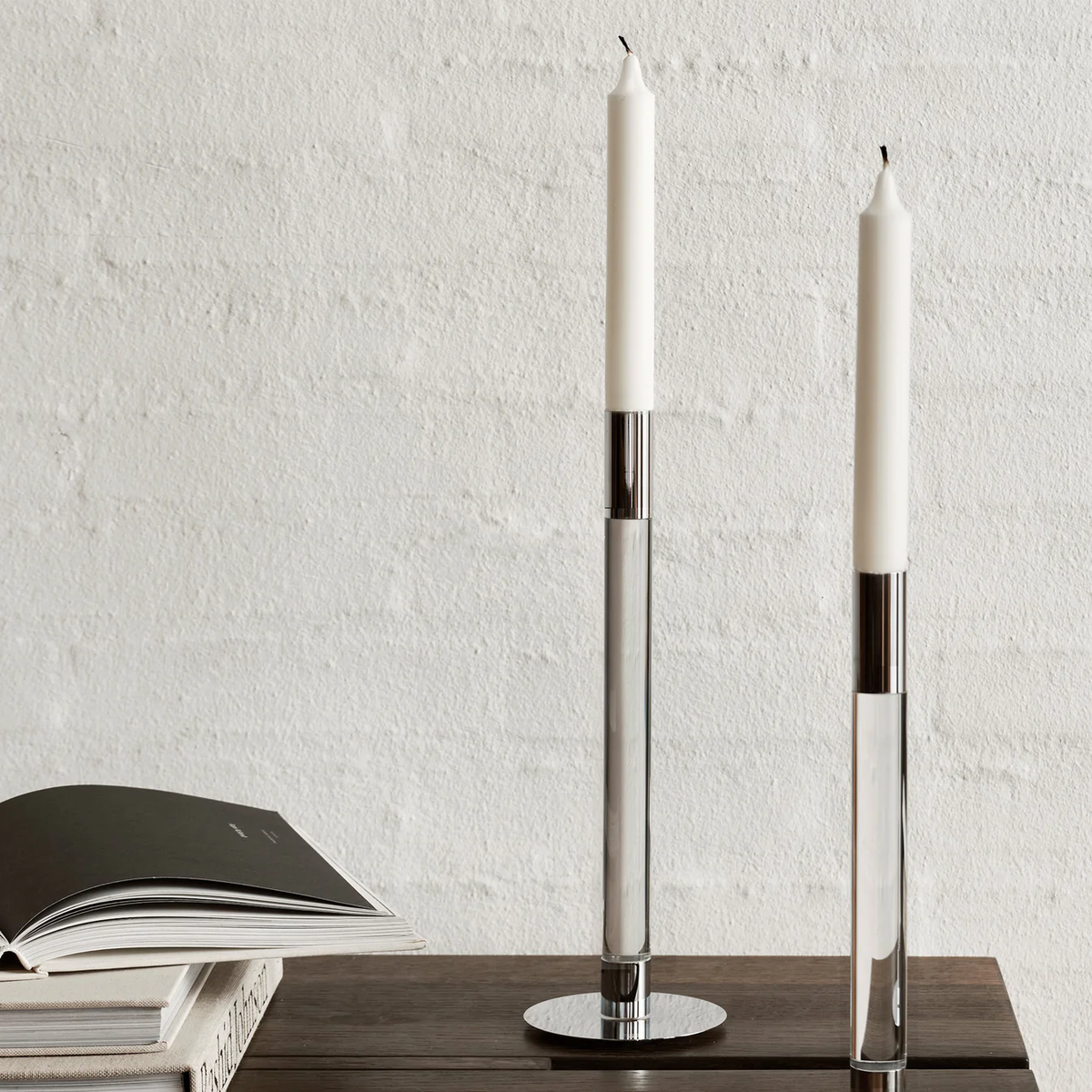 Two tall white taper candles in the Lumiere Candlestick Silver Set of 2 by Orrefors Kosta Boda adorn a wooden surface beside an open book, all set against a textured white wall for modern appeal.