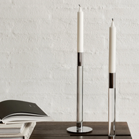 Two tall white taper candles in the Lumiere Candlestick Silver Set of 2 by Orrefors Kosta Boda adorn a wooden surface beside an open book, all set against a textured white wall for modern appeal.