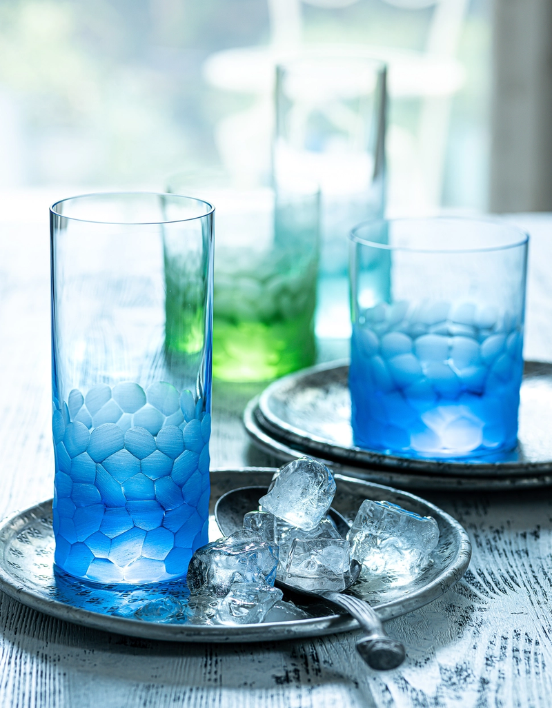 Two silver trays hold Pebbles Highball 400ml glasses by Moser in blue and green, crafted from lead-free Czech crystal, with ice cubes and metal spoons, arranged on a white wooden surface with a softly blurred background.