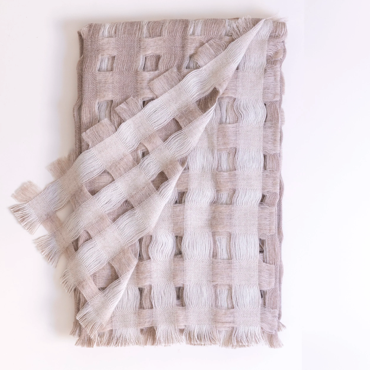 The Dove & Donkey Reversible Space Between Us Throw in Oat & Cloud features a square, open-weave pattern in beige and cream tones with fringed edges, and is shown neatly folded on a white background.