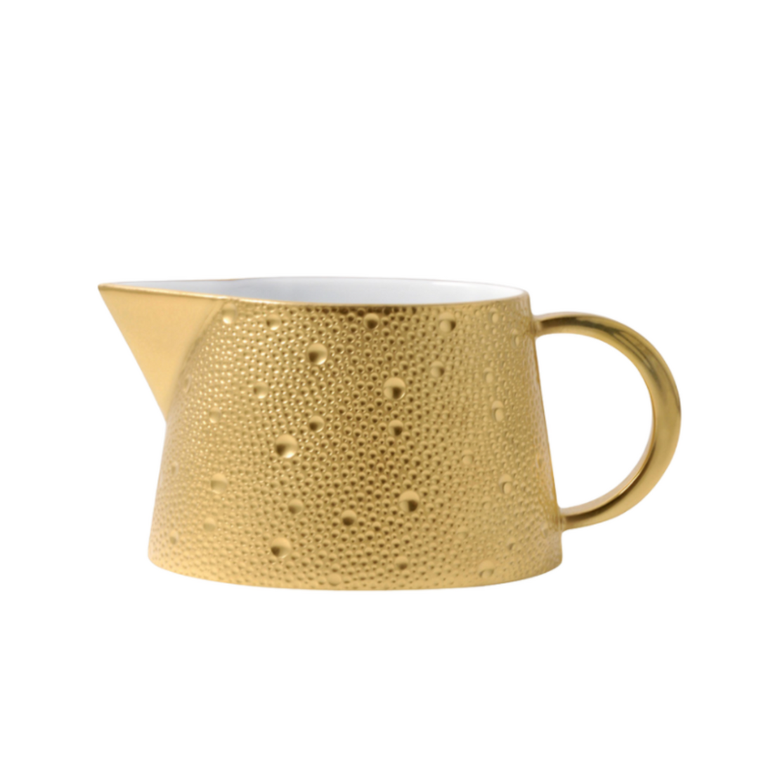 A small golden creamer from Bernardaud's Ecume Dinnerware Gold collection, featuring a textured surface, spout, rounded handle, and white interior—perfect to complement contemporary or Bernardaud tableware.