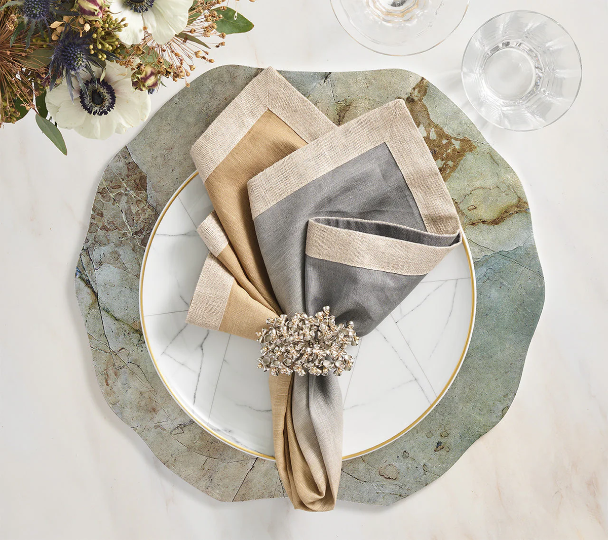 A table setting featuring a green and white marble charger, a gold-rimmed white plate, beige and gray linen napkins with the Kim Seybert Lucent Napkin Ring in Silver & Gold (Set of 4), plus a glass of water—ideal for modern tablescapes.