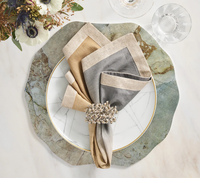 A table setting featuring a green and white marble charger, a gold-rimmed white plate, beige and gray linen napkins with the Kim Seybert Lucent Napkin Ring in Silver & Gold (Set of 4), plus a glass of water—ideal for modern tablescapes.