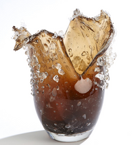 A glass of dark soda shatters, sending ice and liquid flying against a white background. The airborne fragments evoke the unique form of the Dual Wave Vessel by Global Views/Surya at the moment of impact.