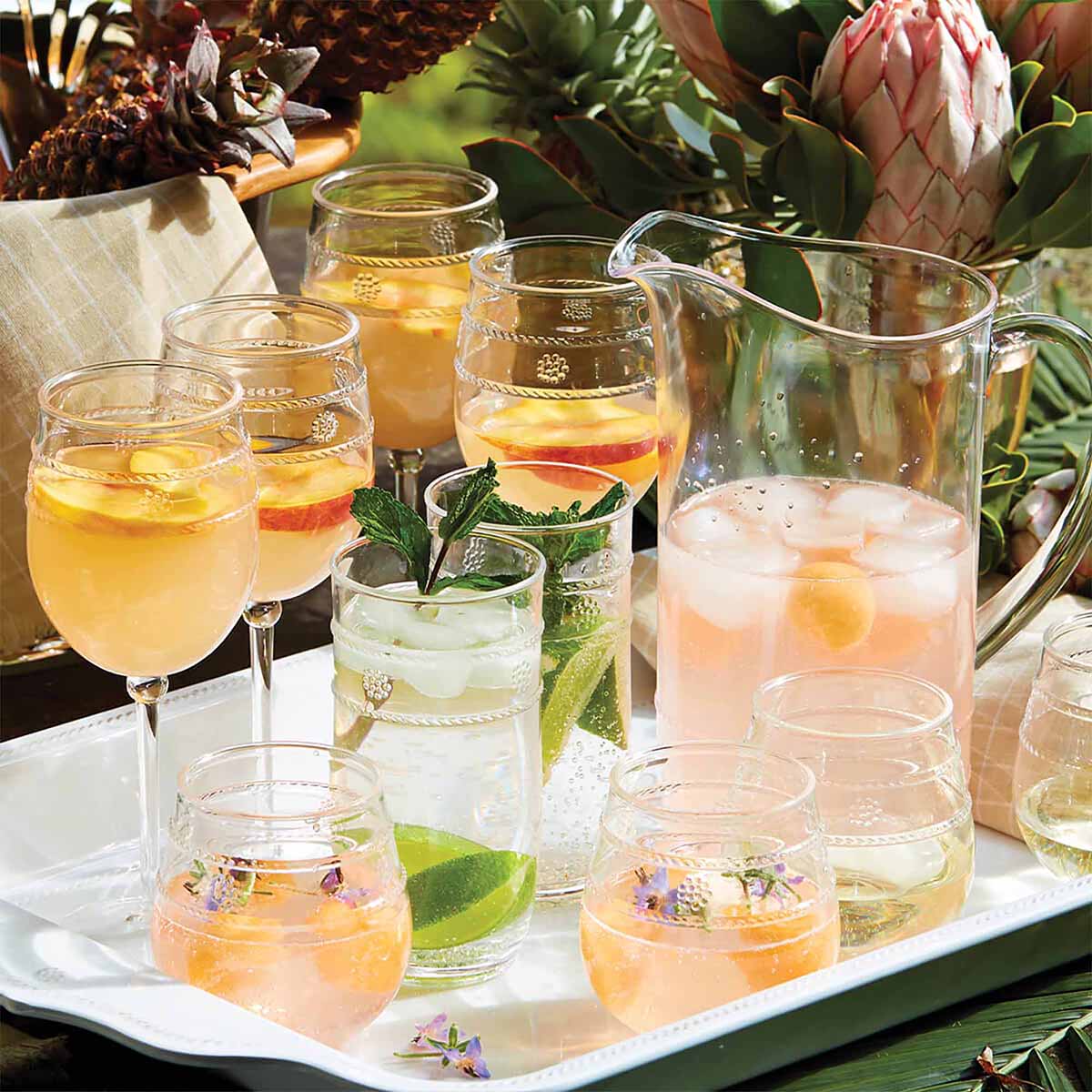 A tray holds the Juliska Isabella Acrylic Pitcher with a pink drink, lemon and peach slices, plus glasses garnished with herbs, citrus, and edible flowers—perfect for outdoor entertaining amid tropical decor.