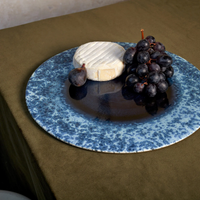 A L'Objet Terra Platter Charger Indigo displays a round of soft white cheese and dark purple grapes, set atop an olive green tablecloth with elegant porcelain placemats beneath.