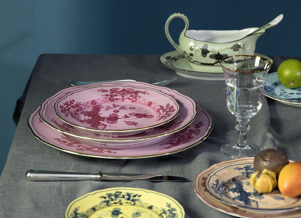 A table set with Ginori 1735 Oriente Italiano Dinnerware Azalea plates, a matching gravy boat, silver cutlery, a glass of water, and assorted fruits on a gray tablecloth.
