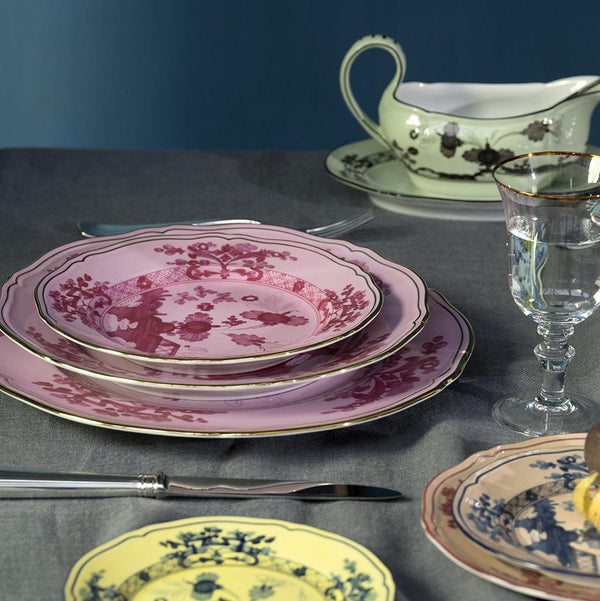 A table set with Ginori 1735 Oriente Italiano Porpora plates in pink, yellow, and blue floral patterns, a glass, silver cutlery, and a green gravy boat on a gray tablecloth against a dark blue background.