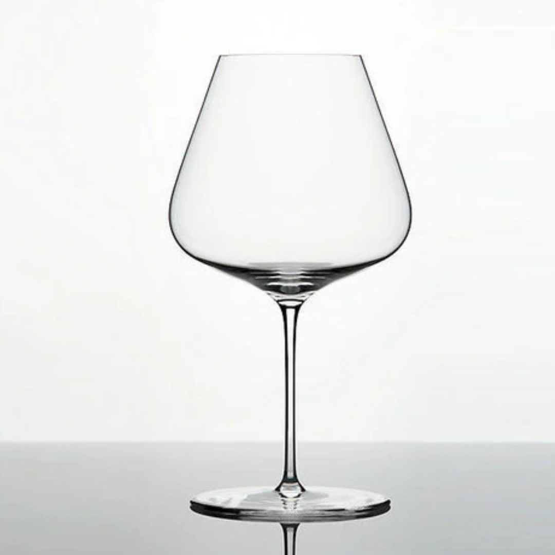 A clear, lead-free Grand Cru Selections / Zalto Burgundy Glass with a wide bowl and thin stem rests on a reflective surface against a plain white background.