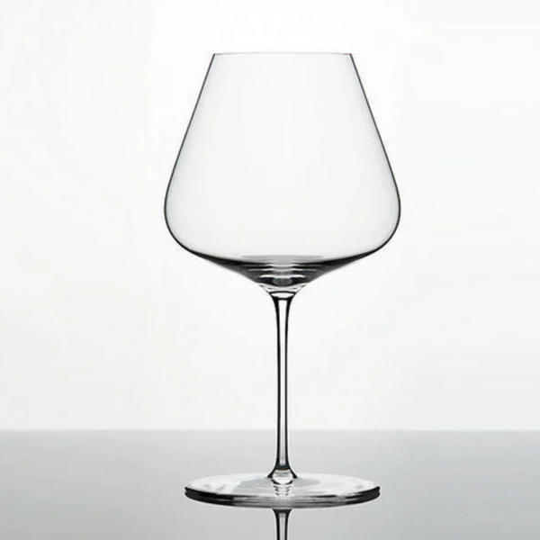 A clear, lead-free Grand Cru Selections / Zalto Burgundy Glass with a wide bowl and thin stem rests on a reflective surface against a plain white background.