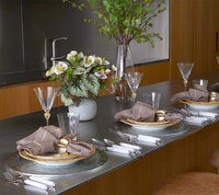 A modern dining setup features gray napkins with gold rings, glass plates, cutlery on a metal countertop, and Kim Seybert’s Daphne Drinkware Set of 4 - Clear & Gold. A vase of flowers completes this elegant look.