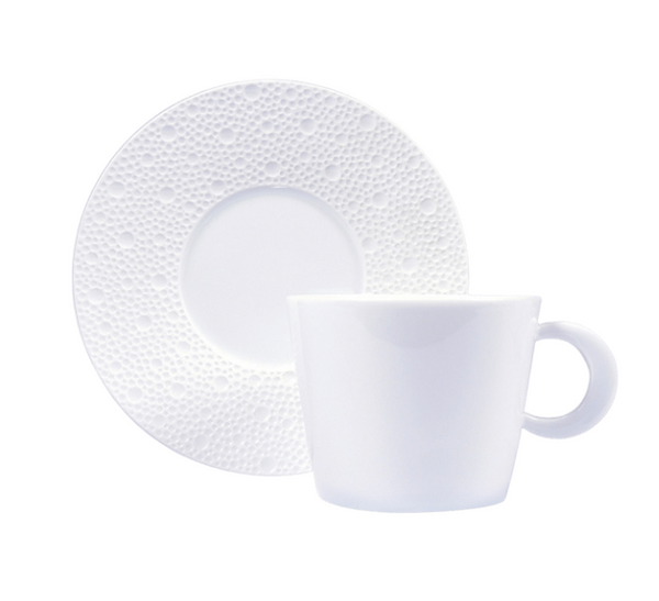 The Ecume White Tea Cup & Saucer by Bernardaud features a simple white ceramic cup with handle and a matching saucer adorned with a textured raised dot pattern around the rim, reflecting minimalist design.