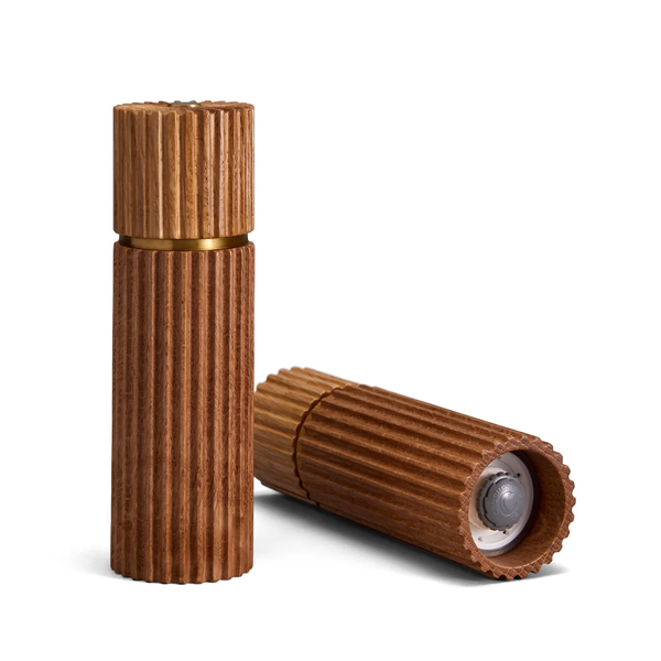 The L'Objet Ionic Salt & Pepper Mill Set Natural Oak features two European oak mills—one upright with a closed lid, the other horizontal and open to reveal a clear inner compartment and metal grinder—for refined seasoning.